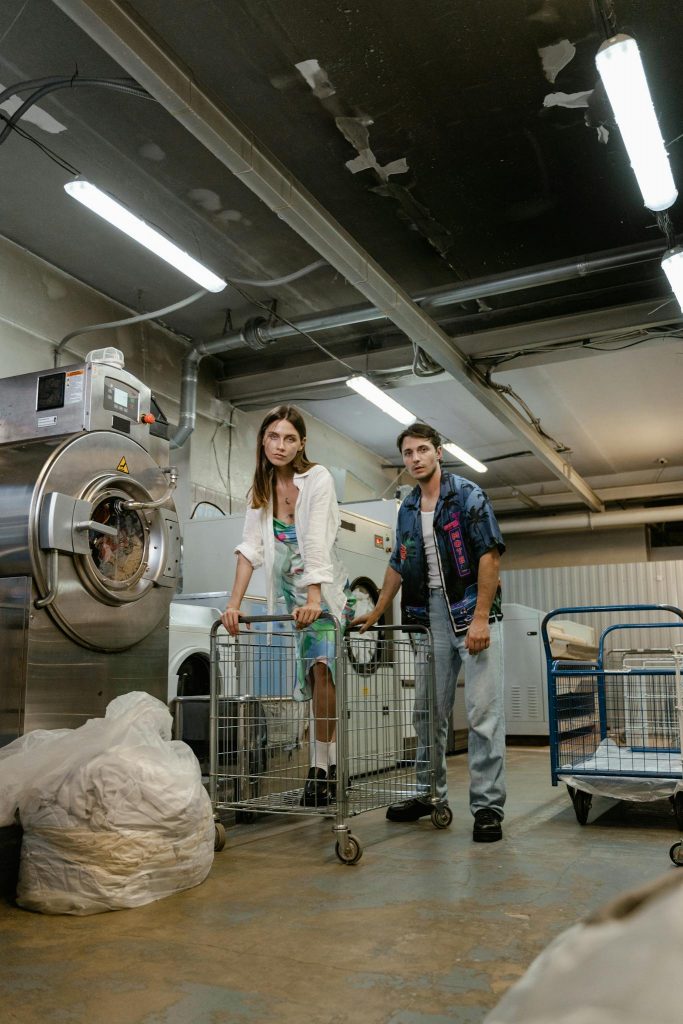 Two young adults standing in a laundromat with industrial washing machines, looking at the camera.
